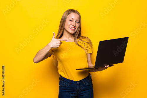 Portrait of a cheerful young casual woman using laptop computer, thumbs up standing isolated over yellow background