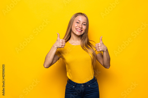 Happy brunette woman showing thumbs up and looking at the camera over yellow background