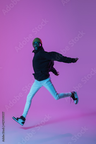 Young stylish man in modern street style outfit isolated on gradient background in neon light. African-american fashionable model in look book  musician performing.