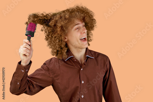 A long-haired curly-haired guy in a brown shirt on a colored background uses a wooden comb. Emotions before a haircut in a hairdresser. Pain from combing