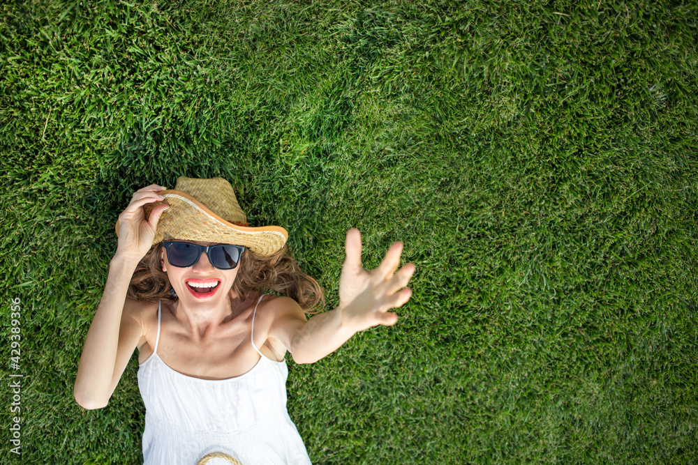 Woman wearing hat and sunglasses and lying on green grass, top view	