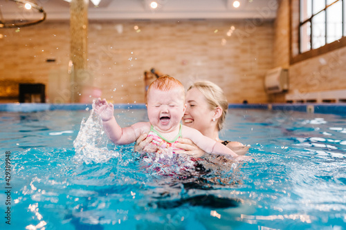 Young mother  swimming instructor and happy little girl in paddling pool. Teaches infant child to swim. Enjoy first day of swimming in water. Mom holds hand child preparing for diving. doing exercises