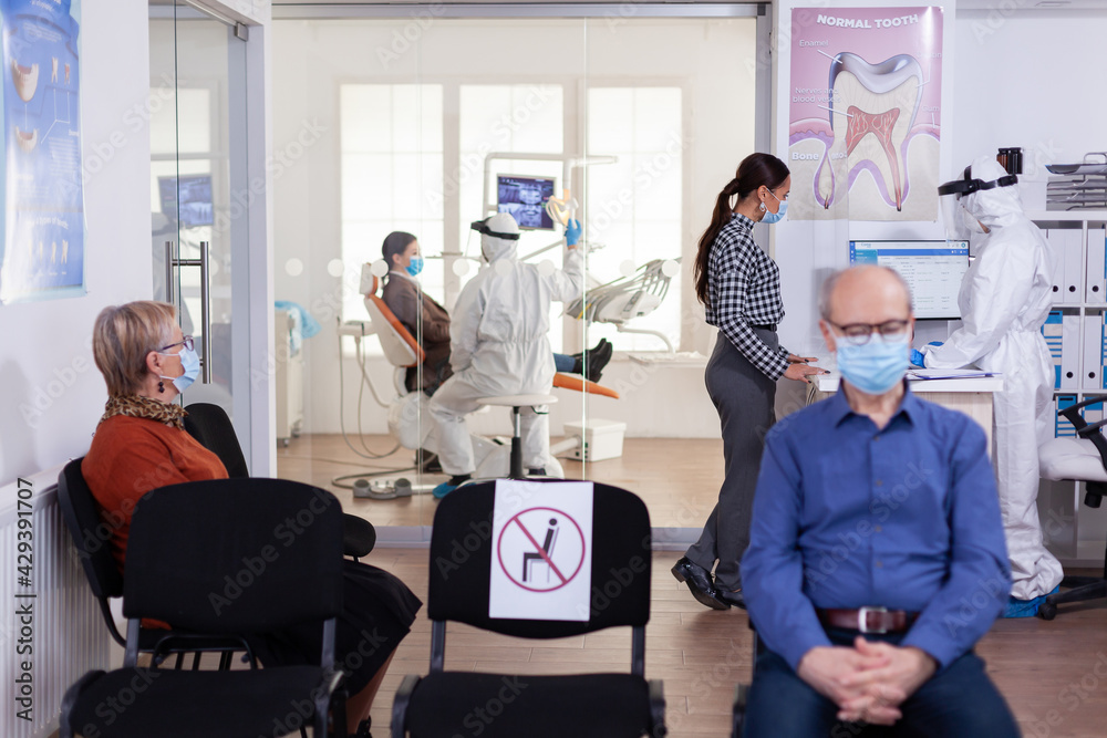 Obraz premium Dentist nurse dressed in ppe suit with face shiled discussing with patient in stomatology waiting room. People keeping social distancing as prevention during coronavirus outbreak.