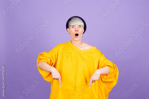 Stylish european woman on purple background pointing down with fingers showing advertisement, surprised face and open mouth