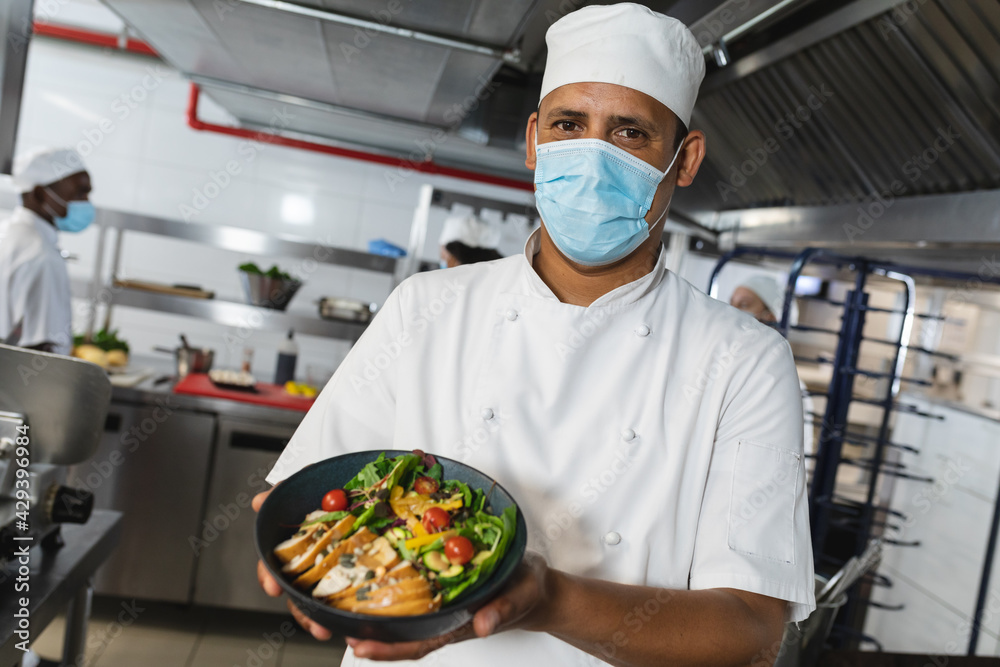 Portrait of mixed race male professional chef with finished dish wearing face mask Portrait of mixed race male professional chef with finished dish wearing face mask