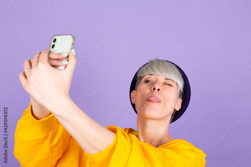 Stylish european woman on purple background playful taking selfie on mobile phone happy emotions positive Stylish european woman on purple background playful taking selfie on mobile phone happy emotions positive