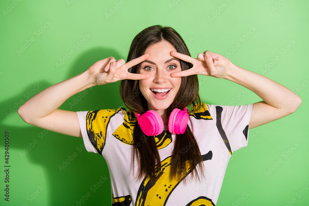 Photo of young happy excited cheerful girl in headphones showing v-sign on eyes isolated on green color background Photo of young happy excited cheerful girl in headphones showing v-sign on eyes isolated on green color background