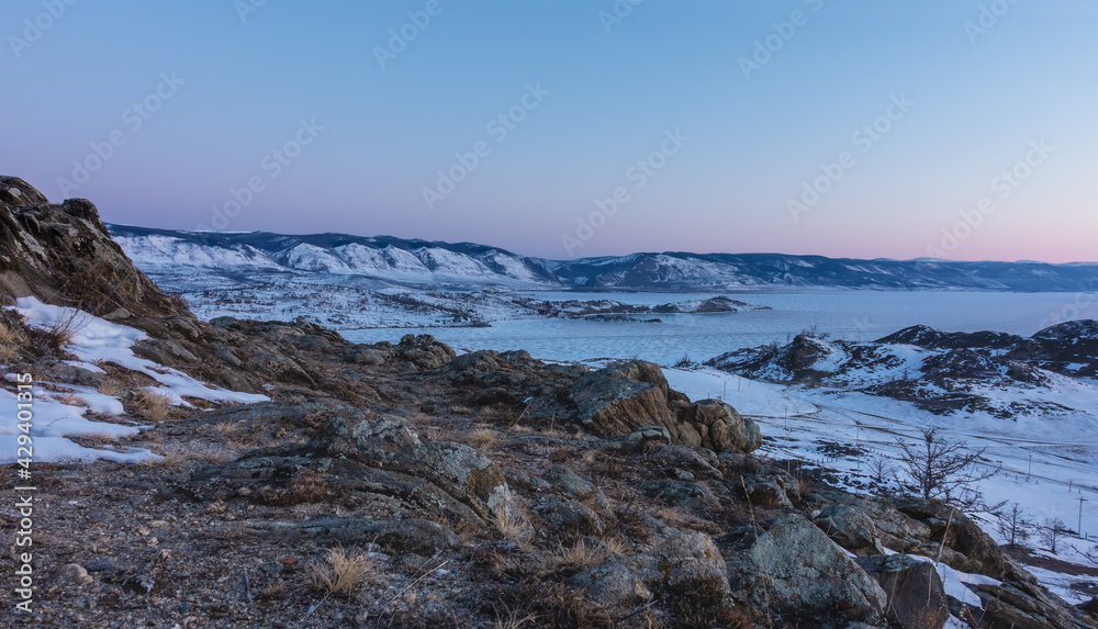 The frozen lake and the rocks around are covered with snow. In the foreground there are picturesque stones, dry grass on the ground. The morning sky is pinkish. Bakal