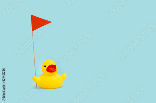 Obraz na plátně Pato de goma amarillo sosteniendo una bandera roja sobre un fondo celeste pastel liso y aislado