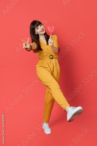 Young beautiful brunette woman wearing stylish ocher suit, holding lollipop, smiling with happy face and showing her leg at the camera, doing victory sign over red background. © sofiko14