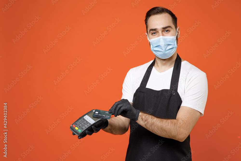 Young man barista barman employee in apron white tshirt face mask coronavirus covid19 quarantine work in shop uses wireless bank payment terminal process acquire payment isolated on orange background Young man barista barman employee in apron white tshirt face mask coronavirus covid19 quarantine work in shop uses wireless bank payment terminal process acquire payment isolated on orange background