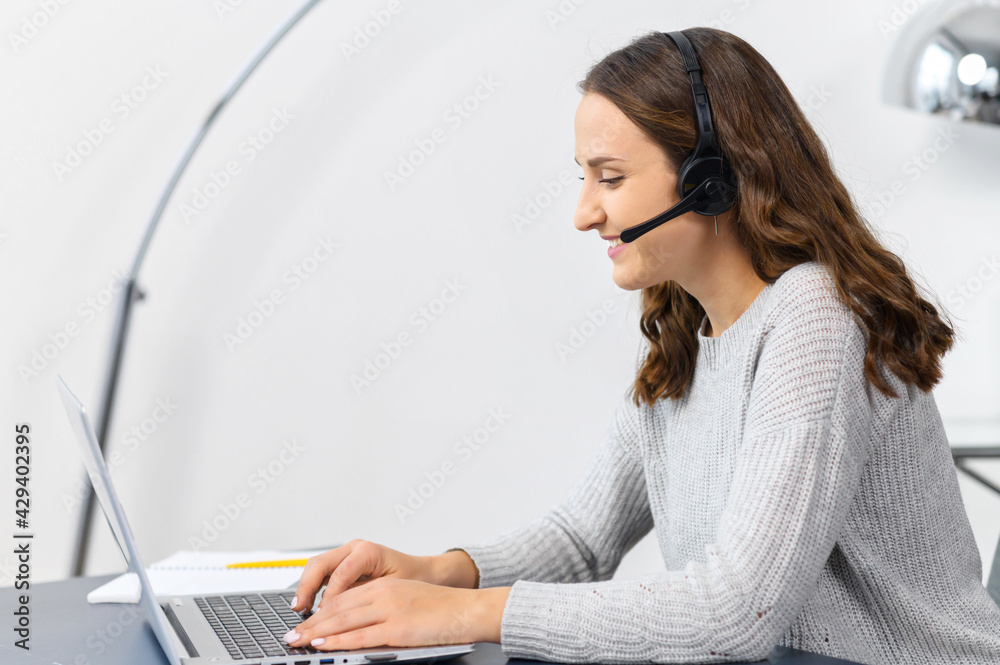 Young woman wearing wireless headset using a laptop computer for communication with customers or colleagues sitting at the desk in contemporary office. Female support worker talks online Young woman wearing wireless headset using a laptop computer for communication with customers or colleagues sitting at the desk in contemporary office. Female support worker talks online