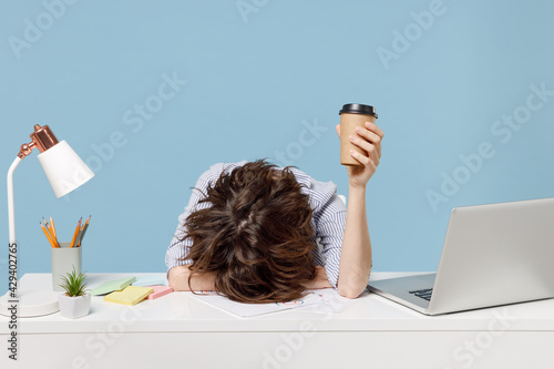 Young tired exhausted secretary employee business woman in casual shirt sit work sleep laid her head down on white office desk with pc laptop hold cup coffee isolated on pastel blue background studio. photo
