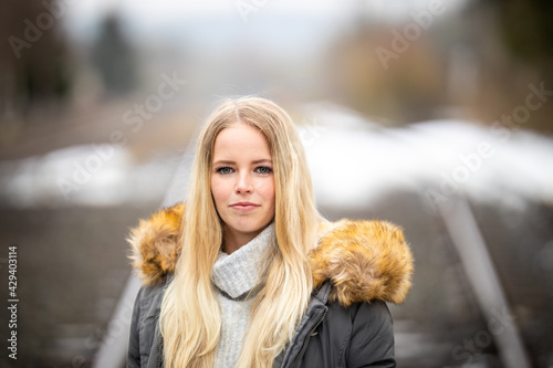 Girl with long blond hair and a gray turtleneck sweater looks seriously at the camera, in the background a wintry track system in the blur..