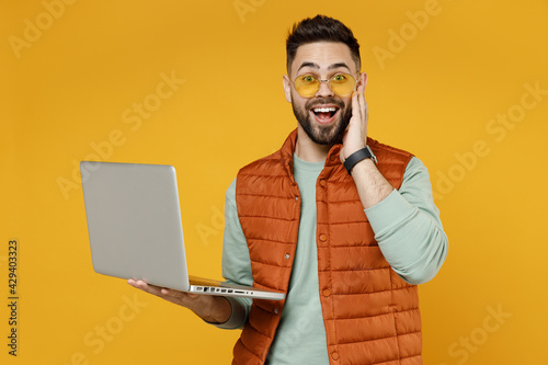 Young freelancer surprised excited caucasian man 20s wearing orange vest mint sweatshirt glasses using laptop pc computer chat online browsing internet hold face isolated on yellow background studio. photo