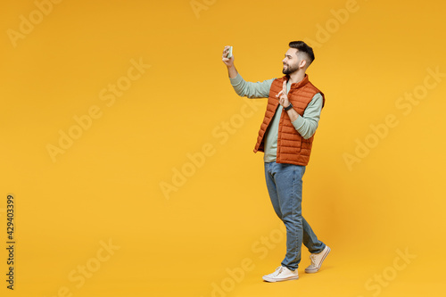 Full length young smiling friendly man in orange vest mint sweatshirt glasses do selfie shot on mobile phone show victory v-sign gesture isolated on yellow background studio. People lifestyle concept.