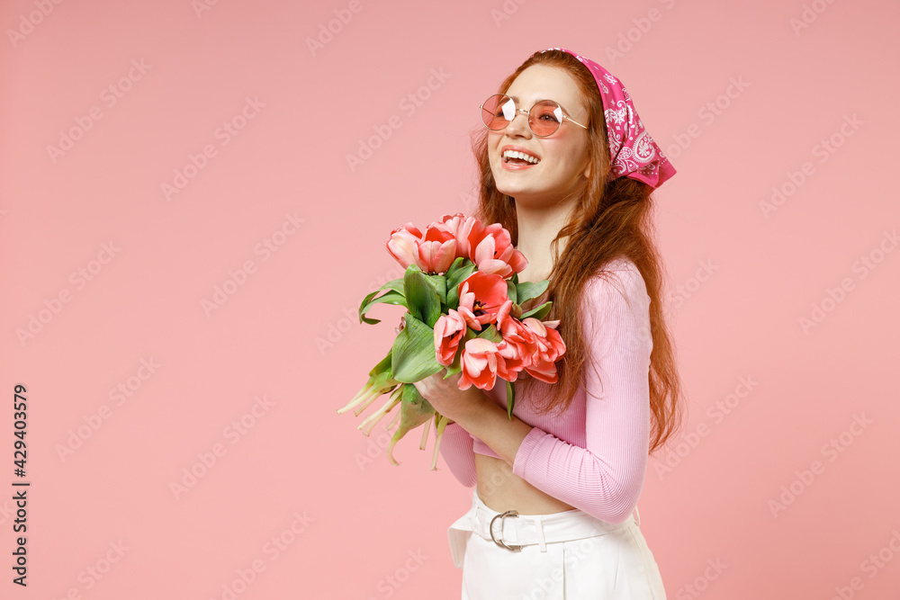 Side view of young smiling fun happy romantic woman 20s wearing rose clothes bandana glasses hold tulips flowers bouquet isolated on pastel pink color background studio portrait Spring season concept Side view of young smiling fun happy romantic woman 20s wearing rose clothes bandana glasses hold tulips flowers bouquet isolated on pastel pink color background studio portrait Spring season concept