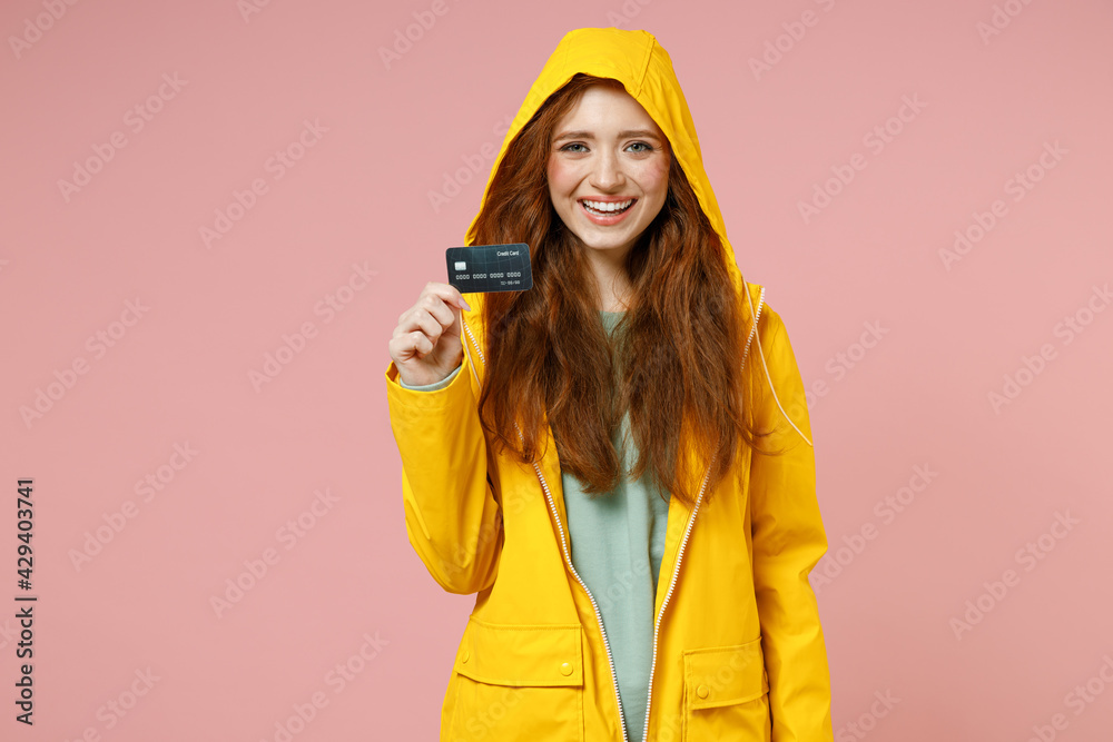 Redhead young smiling woman in yellow waterproof hood raincoat outerwear hold credit bank card isolated on pastel pink color background studio portrait. Outdoors lifestyle fall weather season concept Redhead young smiling woman in yellow waterproof hood raincoat outerwear hold credit bank card isolated on pastel pink color background studio portrait. Outdoors lifestyle fall weather season concept