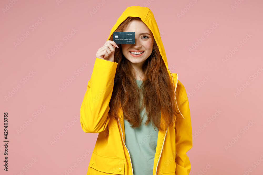 Redhead young cheerful woman in yellow waterproof raincoat hood outerwear cover eye with credit bank card isolated on pastel pink background studio portrait. Lifestyle wet weather fall season concept Redhead young cheerful woman in yellow waterproof raincoat hood outerwear cover eye with credit bank card isolated on pastel pink background studio portrait. Lifestyle wet weather fall season concept