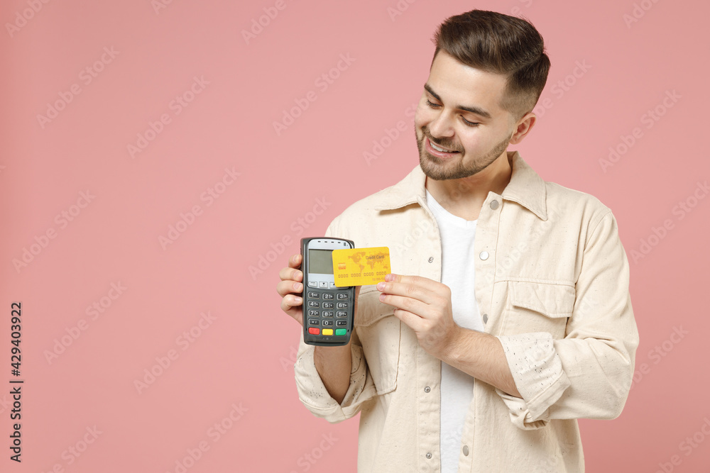 Young smiling caucasian rich man in jacket white t-shirt holding wireless modern bank payment terminal to process and acquire credit card payments isolated on pastel pink background studio portrait. Young smiling caucasian rich man in jacket white t-shirt holding wireless modern bank payment terminal to process and acquire credit card payments isolated on pastel pink background studio portrait.