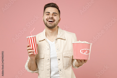 Young smiling laughing cheerful fun man 20s wearing jacket white t-shirt hold popcorn takeaway bucket soda cola paper cup isolated on pastel pink background studio portrait. Lifestyle people concept.
