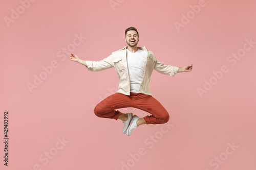 Full length young happy spiritual man 20s in jacket white t-shirt jump hold spreading hands in yoga om gesture relax meditate try to calm down isolated on pastel pink color background studio portrait