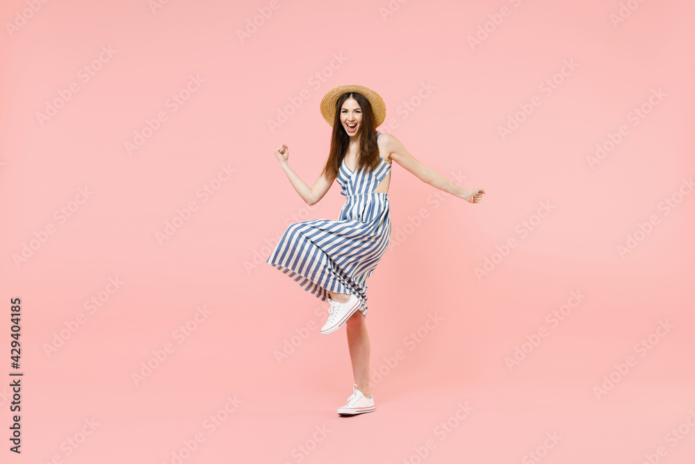 Full length young happy woman in summer clothes striped dress straw hat do winner gesture clench fist with raised up leg isolated on pastel pink background studio portrait. People lifestyle concept.