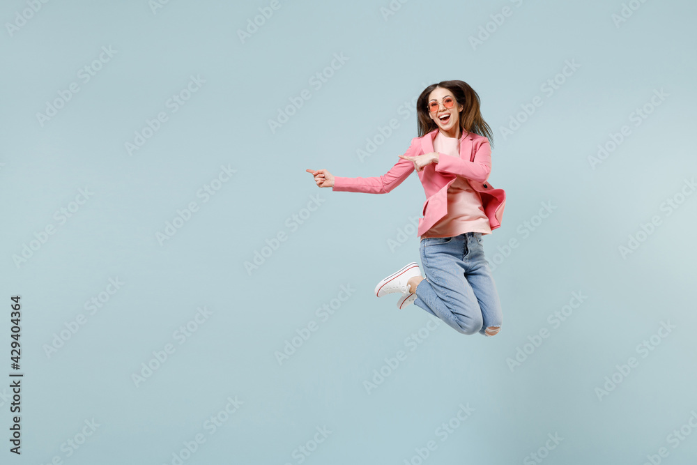Full length young smiling happy trendy woman in pastel pink clothes glasses jump high point index finger aside on workspace area mock up isolated on blue background studio People lifestyle concept