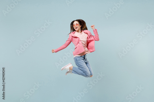 Full length young fun smiling happy caucasian trendy stylish woman in pastel pink clothes glasses jump high do winner gesture clench fist isolated on blue background studio. People lifestyle concept.