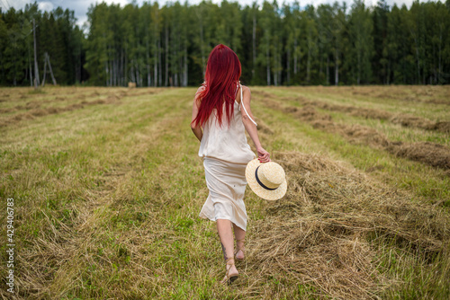 a woman with red hair in a white dress and a straw hat in her hands runs across a meadow field