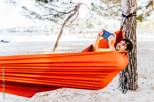 Hispanic man lying in a hammock and playing video games at Palmanova beach. Palma de Mallorca, Spain (Perfect for Copyspace)	