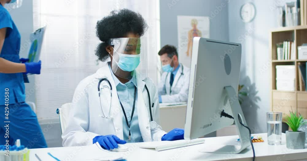 Portrait of beautiful busy African American young woman physician in medical mask and face shield typing on computer searching internet sitting in cabinet in medical center. Medic concept