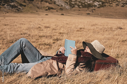 Woman traveler with hat is lying down in a golden grass field and is reading a book.