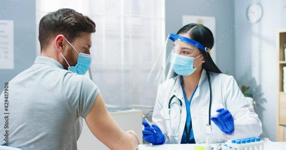 Close up of young Asian pretty female medic physician in medical mask and face shield filling syringe with covid-19 vaccine doing injection in hospital to handsome Caucasian male patient. Vaccination Close up of young Asian pretty female medic physician in medical mask and face shield filling syringe with covid-19 vaccine doing injection in hospital to handsome Caucasian male patient. Vaccination