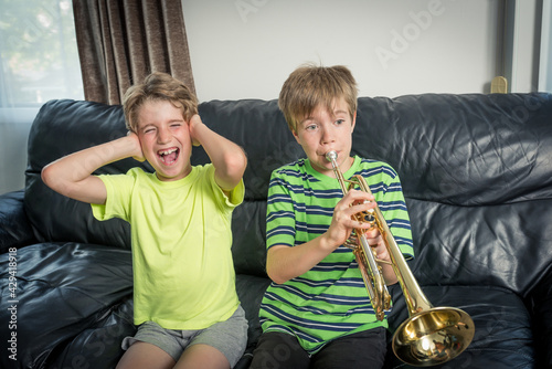 Two kids sitting on a sofa.
One is playing a trumpet and the other child annoyed is covering his ears with his hands photo