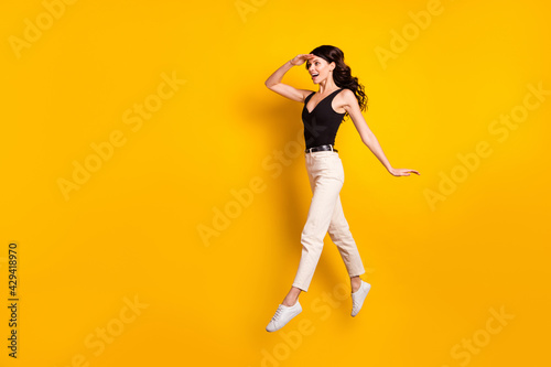 Full length body size photo of funny woman jumping keeping hand near forehead looking far isolated vivid yellow color background