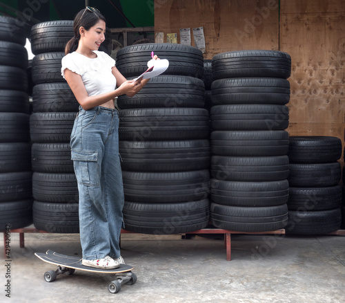 Young beautiful woman standing on surf skateboard at pile of used dirty rubber tires in background. Asian female stays outdoor smiling check inventory of her second hand tyres recycling business store photo