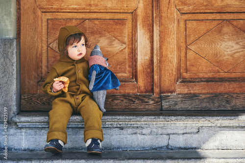 Outdoor portrait of adorable toddler girl eating croissant, playing with vintage teddy bear, wearing warm jumpsuit, fashion for little children