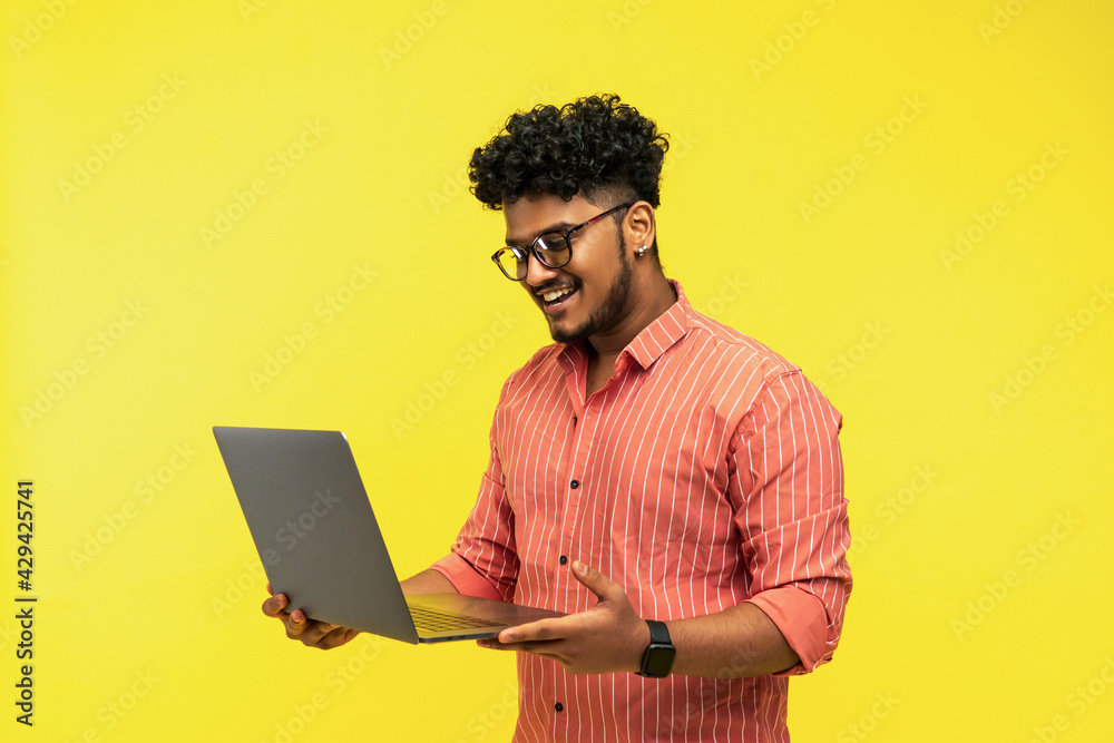 Smiling Indian man with laptop in hands isolated on yellow background, smiling looking at computer, place for text