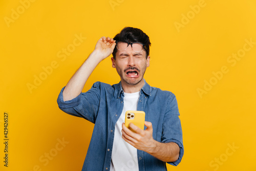 Upset crying caucasian guy in denim shirt holding smartphone in his hand, got a bad news or message, raised his glasses, unhappy facial expression, stands on isolated orange background