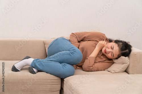 Unhappy Asian woman sitting on the sofa and holding on stomach suffering. Abdominal pain that comes from menstruation, diarrhea, or indigestion. Sickness and healthcare concept