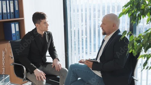 Portrait of two businessman sitting and chatting with office background. Casual looking men in suits discussing oportunities and upcoming projects. Concept of interview, deal. photo