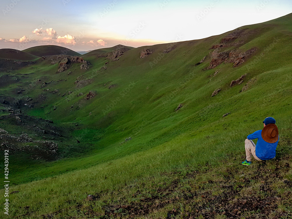 A man sitting on a stone on a green hill in Xilinhot, Inner Mongolia and enjoys a sunset. He is wearing a cowboy hat. The sun starts to set behind the hills, turning the sky orange. Freedom