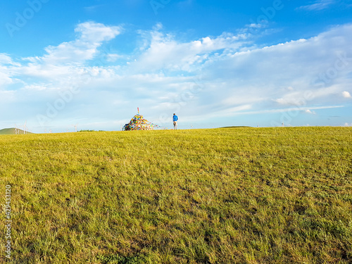 A man standing on a vast pasture in Xilinhot in Inner Mongolia, and admiring a heap of stones (aobao) with colorful prayer flags attached to it. Endless grassland. Blue sky with white clouds. Spirits photo