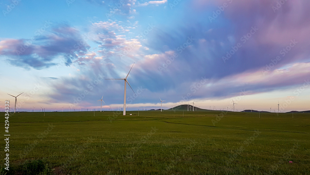A panoramic view on a hilly landscape of Xilinhot in Inner Mongolia. Endless grassland with a few wind turbines in the back. The sun starts to set, coloring the sky pink. Thick, rainy clouds.