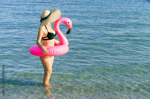 Summer fun. Young sexy woman in straw hat, bikini swimsuit, sunglasses with pink inflatable flamingo in blue ocean water for sea beach background. Spa and sunbathing outdoor.