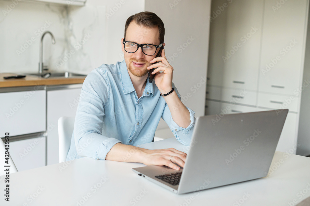 Work from home. Clever and positive young businessman talks on the phone using laptop computer for remote work from home, a guy in casual shirt sits at the table in the kitchen Work from home. Clever and positive young businessman talks on the phone using laptop computer for remote work from home, a guy in casual shirt sits at the table in the kitchen