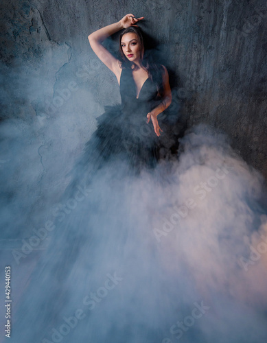 Front view of beautiful brunette in a black dress all in smoke posing on the background of the wall. Concept of lush evening dresses