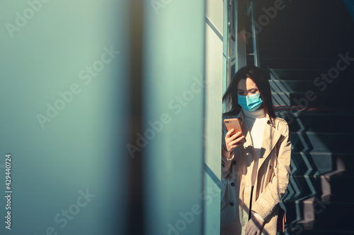Girl with medical mask is using her smartphone in a modern building during pandemic of Coronavirus Covid-19 © danieleorsi