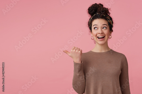 Portrait of attractive with dark curly hair bun. Wearing headband, earrings and brown sweater. Has make up. Watching and pointing thumb to the left at copy space, isolated over pastel pink background